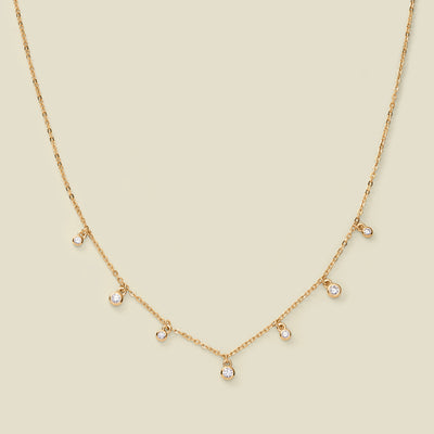 A dainty gold chain necklace with bezel-set clear, round gemstones stationed along it, set against a solid beige background.