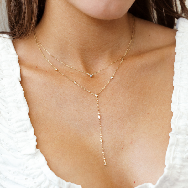 A close up of a person wearing two layered gold necklaces, one with a single stationary clear stone in the center and the other a Y-shape with multiple bezel-set, clear gemstones stationed along it.