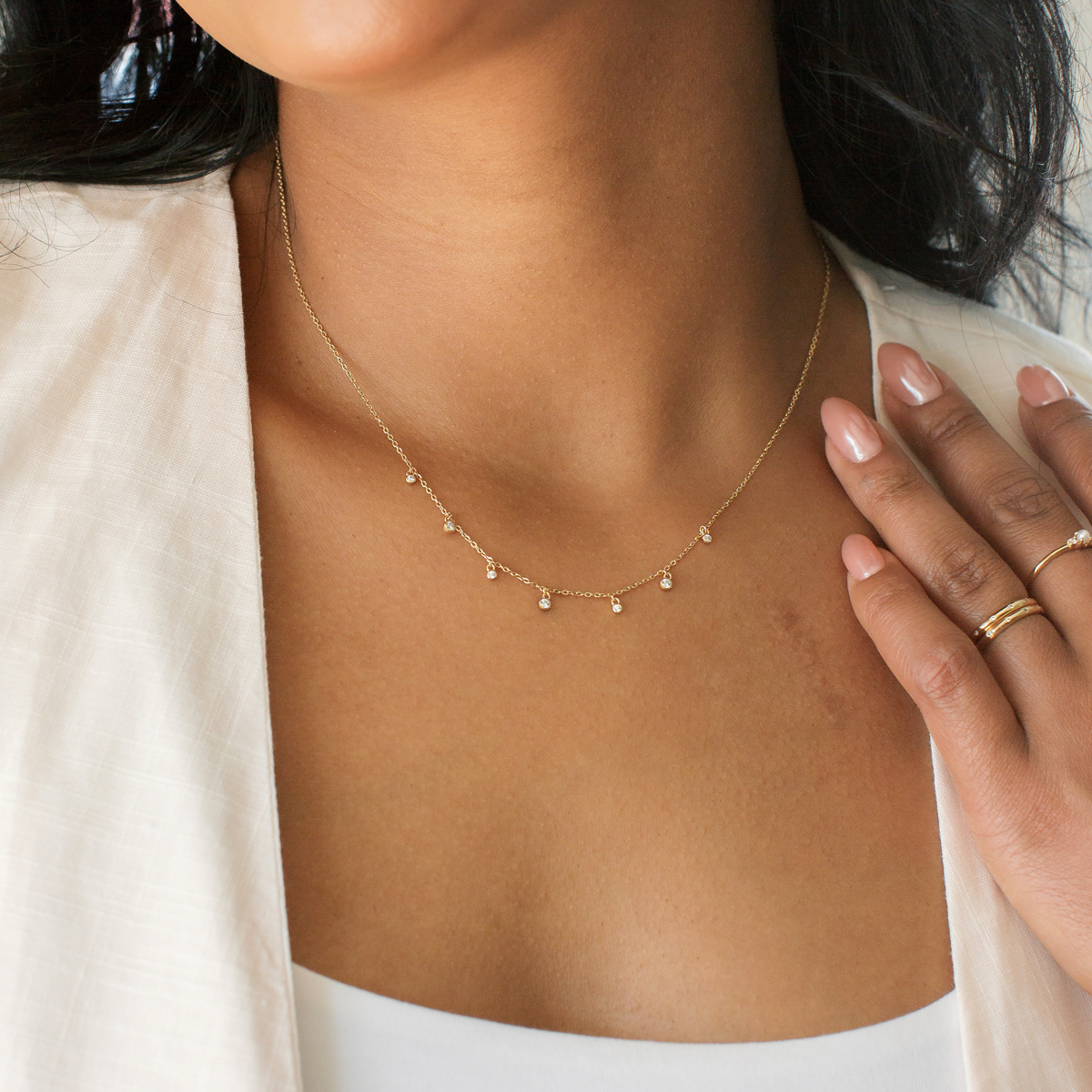 Close-up of a person wearing a delicate gold necklace with dangling, round, clear gemstones stationed along it, paired with a white linen blouse.