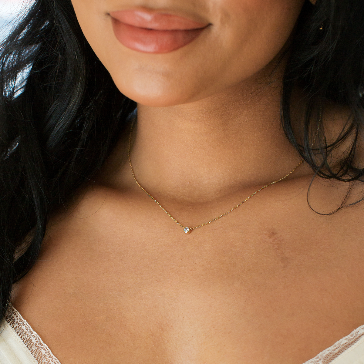 Close-up of a woman wearing a delicate gold chain necklace with a singular bezel-set, round, clear gemstone in the center.