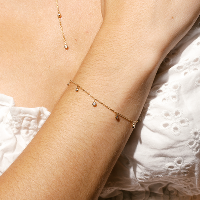 A close-up of a person's wrist wearing a dainty gold chain bracelet with stationary clear, round bezel-set gemstones dangling off of it, with a white lace top.