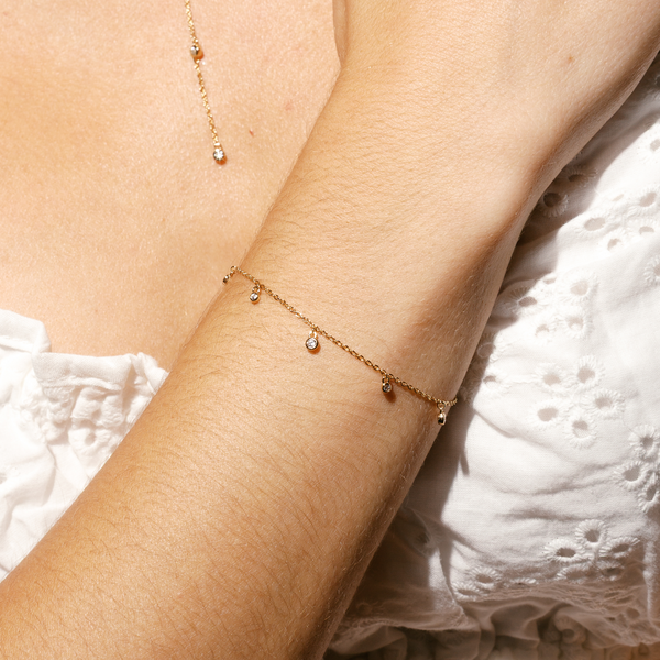 A close-up of a person's wrist wearing a dainty gold chain bracelet with stationary clear, round bezel-set gemstones dangling off of it, with a white lace top.