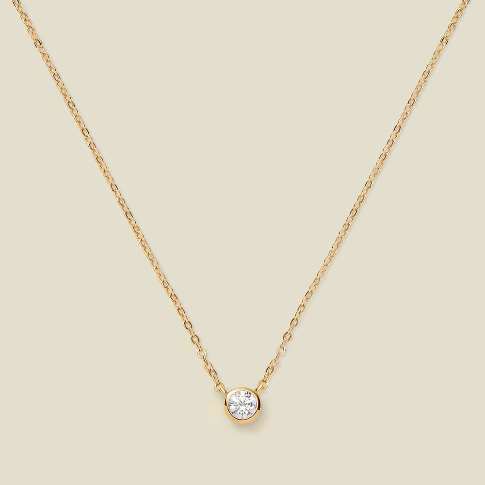 A delicate gold chain necklace with a small round bezel-set clear gemstone pendant, displayed against a solid light beige background.