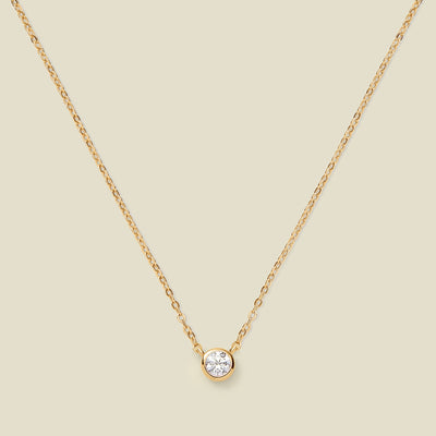 A delicate gold chain necklace with a small round bezel-set clear gemstone pendant, displayed against a solid light beige background.
