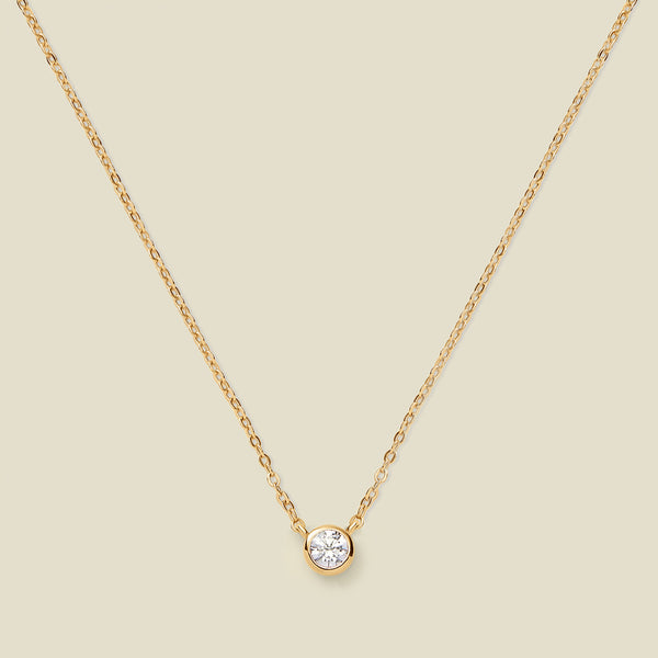 A delicate gold chain necklace with a small round bezel-set clear gemstone pendant, displayed against a solid light beige background.