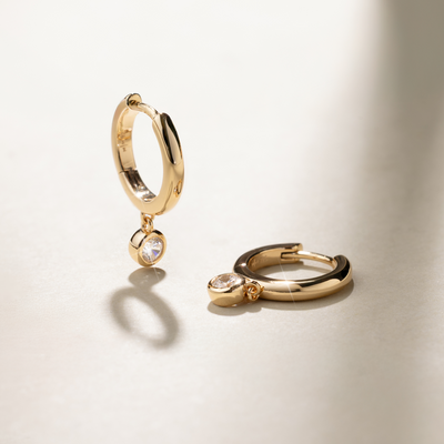A pair of polished gold hoop earrings, each with a singular round bezel-set clear gemstone hanging from the bottom, set against a beige background with natural shadows.