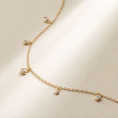 A gold chain bracelet with round, clear bezel-set gemstones dangling off of it, set against a neutral background with natural shadows.