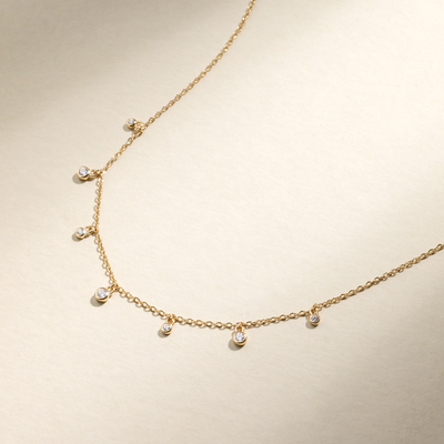 A dainty gold chain necklace with seven stationary bezel-set round clear gemstones hanging off of it, set against a beige background with natural shadows.