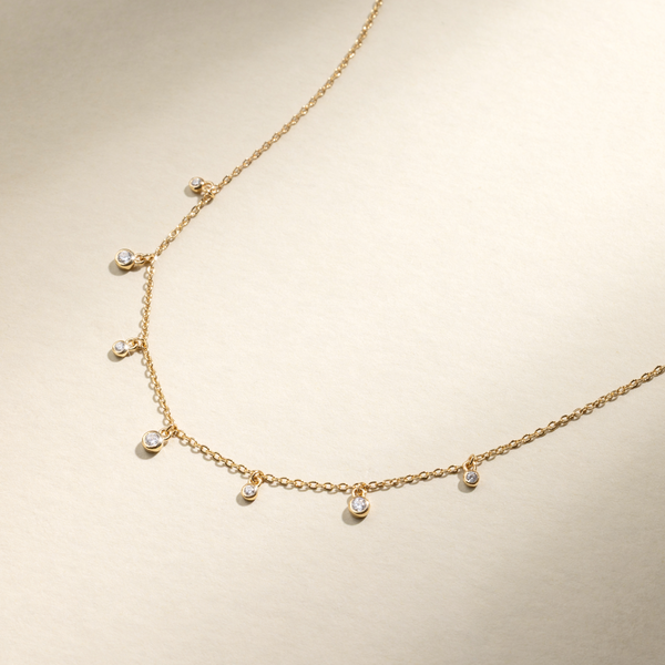 A dainty gold chain necklace with seven stationary bezel-set round clear gemstones hanging off of it, set against a beige background with natural shadows.