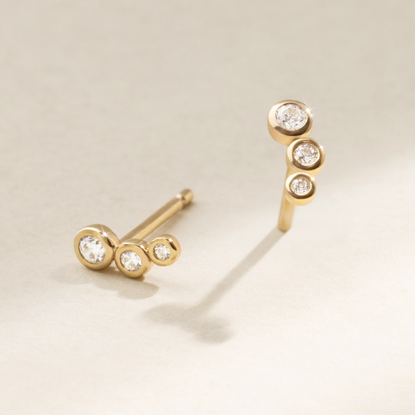 A pair of gold stud earrings, each with three round clear bezel-set gemstones going from largest to smallest in a slight curve, set against a soft beige background with natural shadows.
