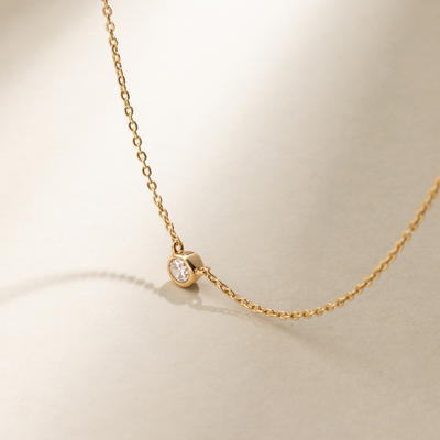 A delicate gold chain necklace with a singular stationary bezel-set round clear gemstone in the center, set against a neutral background with natural shadows.
