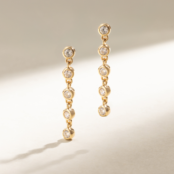 A pair of gold stud earrings with five round, clear bezel-set gemstones dangling vertically, set against a neutral background with natural shadows.