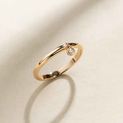 A polished gold ring with a singular bezel-set round clear gemstone hanging from the front, set against a neutral background with natural shadows.