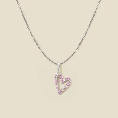 A dainty silver chain necklace with a silver heart charm with different pink colored stones, set against a solid beige background.