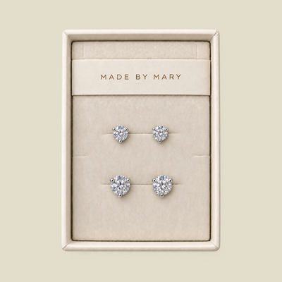 A set of four stud earrings displayed in a cream-colored jewelry box. The earrings feature round clear gemstones in silver settings, with one smaller pair and one larger pair.