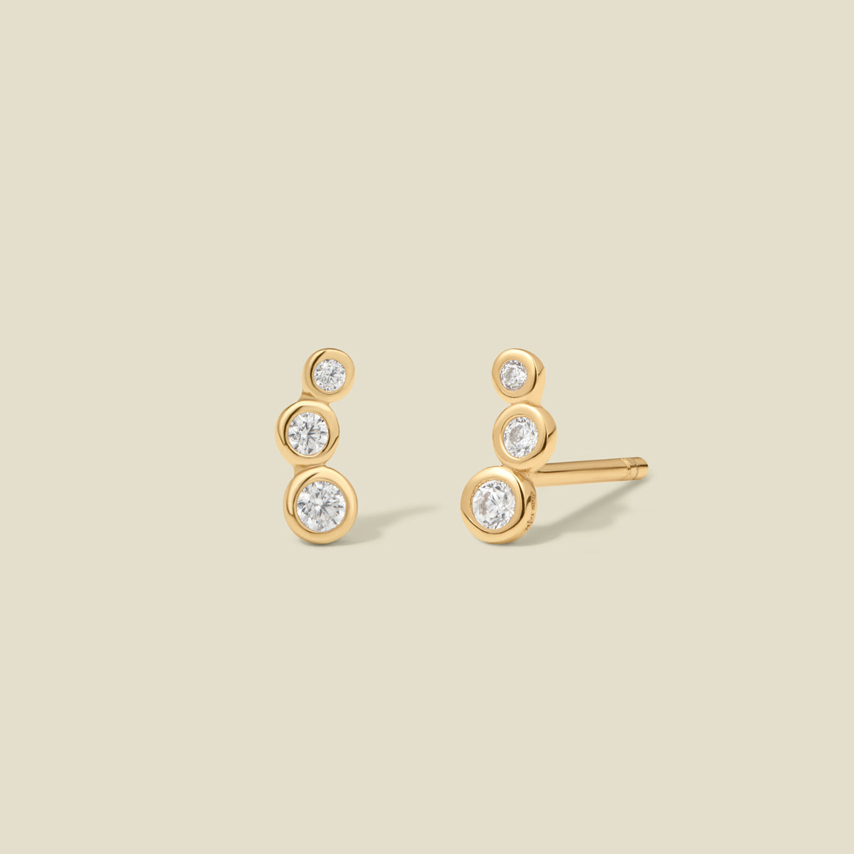 A pair of gold stud earrings with three round, clear gemstones in various sizes curving inward, set against a solid beige background.