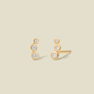 A pair of gold stud earrings with three round, clear gemstones in various sizes curving inward, set against a solid beige background.