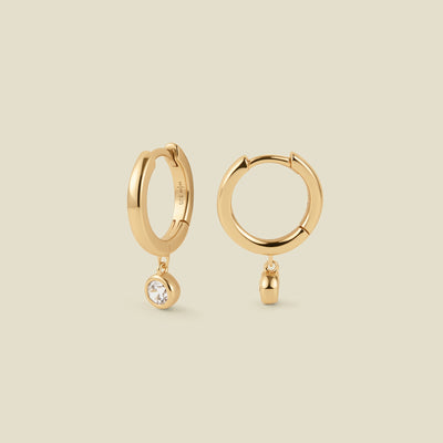 A pair of polished gold hoop earrings, each with a small, round, clear gemstone hanging from the bottom, set against a solid beige background.