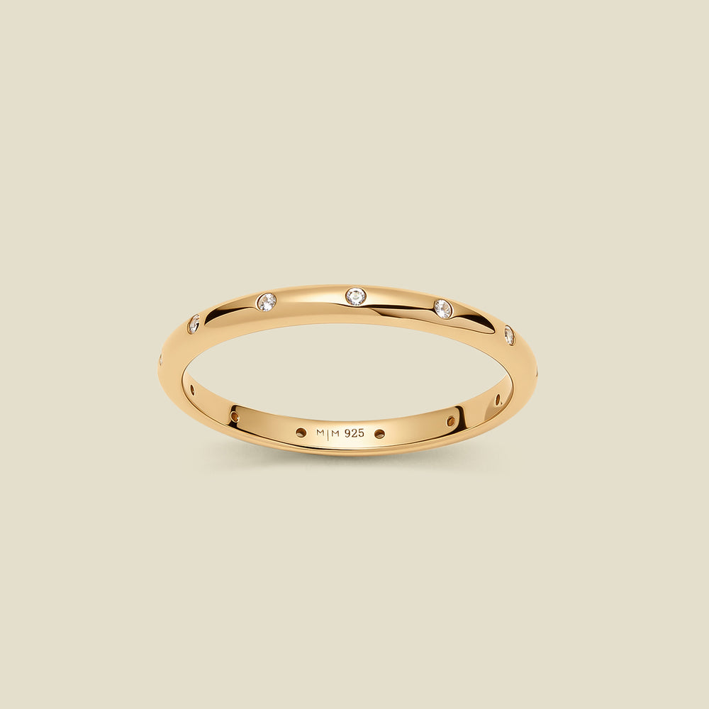 A polished gold ring, inlayed with small, round clear gemstones running along the outside, set against a solid beige background.