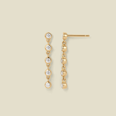 A pair of gold stud earrings with five, round clear gemstones hanging vertically, set against a solid beige background.