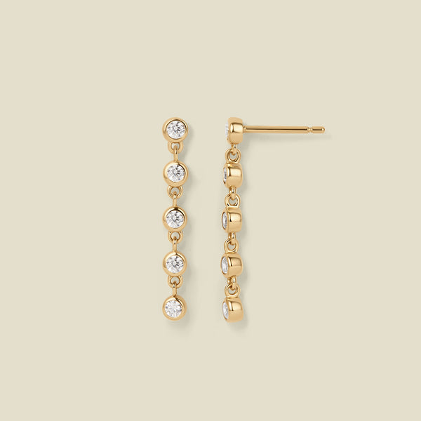 A pair of gold stud earrings with five, round clear gemstones hanging vertically, set against a solid beige background.