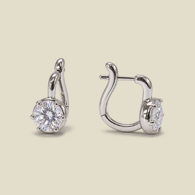 A pair of silver-toned huggie hoop earrings, one shown from the front with a round brilliant-cut clear gemstone and the other from the side, set against a solid light beige background.