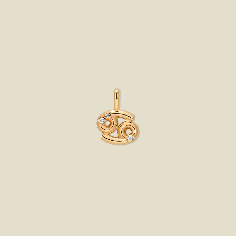 A gold pendant shaped like the Cancer zodiac symbol, featuring three small clear gemstones, centered on a solid light beige background.