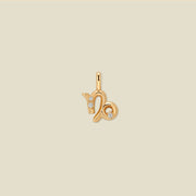 A gold pendant shaped like the Capricorn zodiac symbol, featuring three small clear gemstones, centered on a solid light beige background.