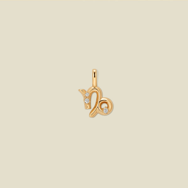 A gold pendant shaped like the Capricorn zodiac symbol, featuring three small clear gemstones, centered on a solid light beige background.