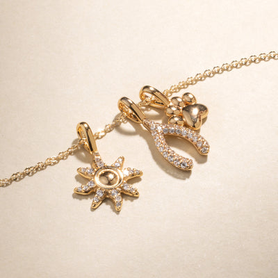A delicate gold chain necklace with a sun charm with clear accent stones, a wishbone charm with clear gemstones, and a polished gold paw print charm against a beige background.