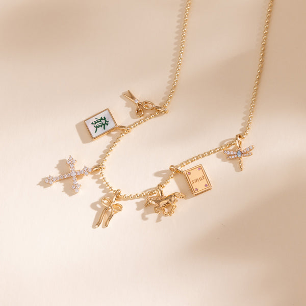 A gold-tone charm necklace with multiple whimsical charms, including a mahjong tile with white inlay and a green character, a cross with clear stones, a small horse, and a book charm, displayed on a warm beige background.