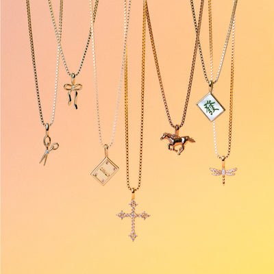 Six delicate chain necklaces with various charms are displayed against a warm peach-to-yellow gradient background. The necklaces feature gold chains with pendants shaped like scissors, a ribbon, a book, a horse, a mahjong tile with green characters, and a dragonfly.