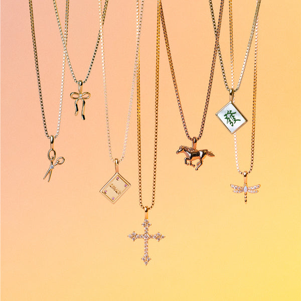 Six delicate chain necklaces with various charms are displayed against a warm peach-to-yellow gradient background. The necklaces feature gold chains with pendants shaped like scissors, a ribbon, a book, a horse, a mahjong tile with green characters, and a dragonfly.