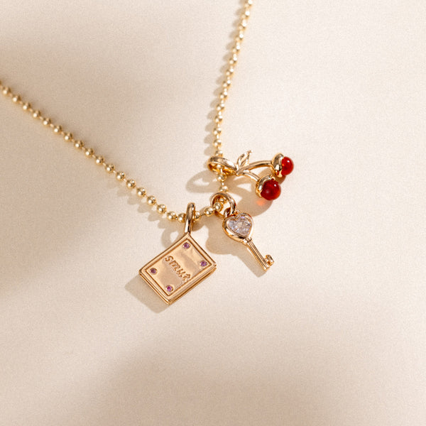 A delicate gold bead chain necklace with three charms, a gold cherry charm with two red agate stones, a gold key charm with a clear, heart shaped stone, and a book charm with the word 'SMUT' and four pink accent stones.