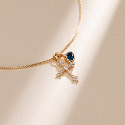 A delicate gold chain necklace featuring a pendant with a small oval blue gemstone and a gold cross adorned with tiny clear crystals, displayed on a warm beige background with soft shadows.