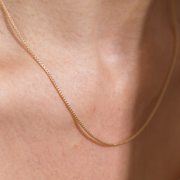 A delicate gold box chain necklace worn on a person's neck, shown in a close-up shot against warm skin tones.