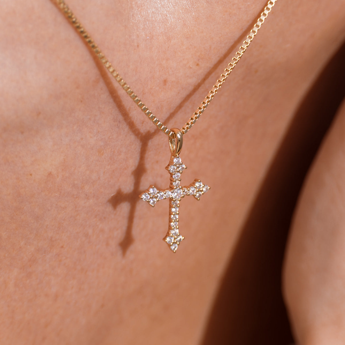 A gold cross pendant necklace adorned with small clear stones, worn on a person's neck against warm skin tones.