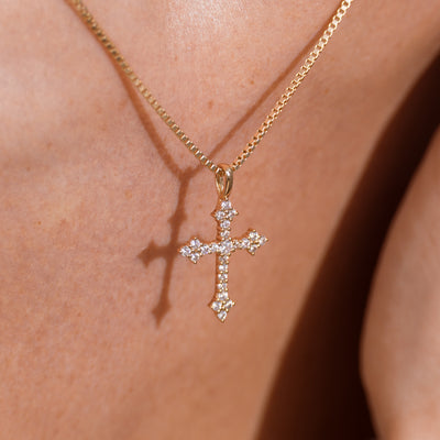 A gold cross pendant necklace adorned with small clear stones, worn on a person's neck against warm skin tones.