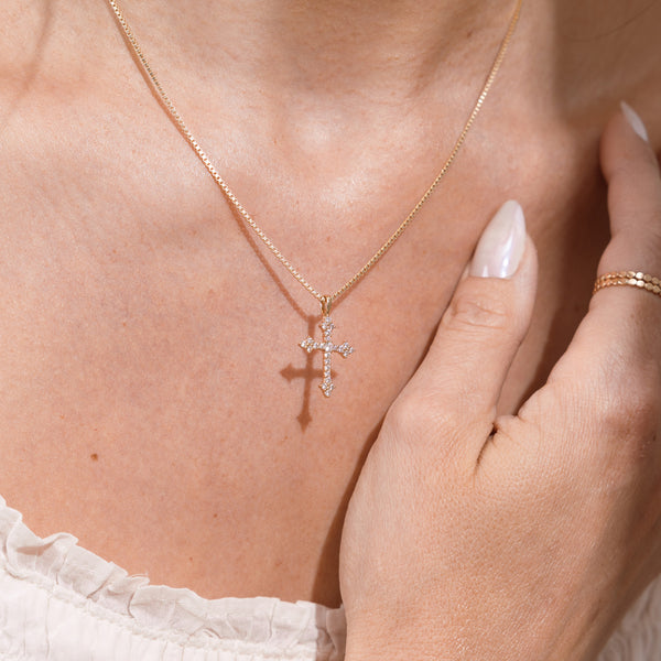 A close-up of a delicate gold cross pendant necklace with small clear stones, worn on a model's neck with a hand resting near the chest.