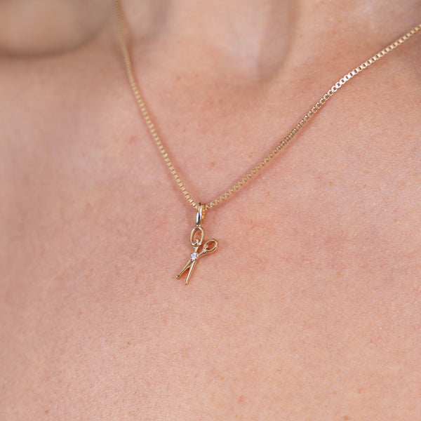 A delicate gold box chain necklace with a small scissors pendant, shown worn on a person's neck.