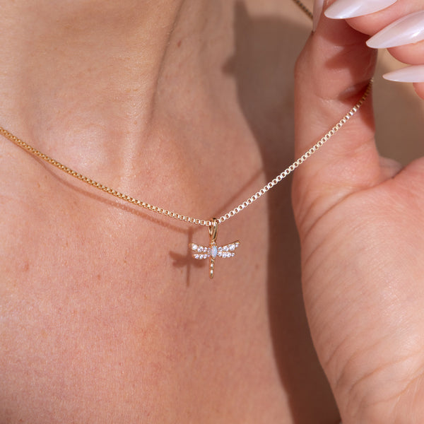A close-up of a delicate gold box chain necklace with a dragonfly pendant, worn on a model's neck with a hand holding the chain nearby.