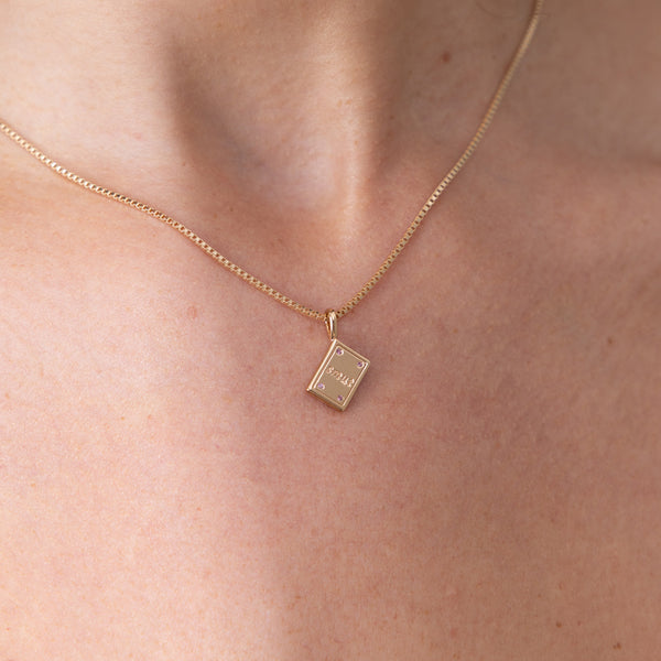 A delicate gold box chain necklace with a small rectangular book pendant engraved with the word 'SMUT', worn on a model's neck.