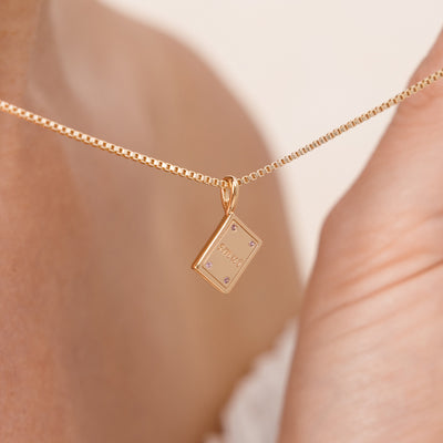 A close-up of a delicate gold box chain necklace worn on a person's neck, featuring a small rectangular book pendant engraved with 'SMUT' and accented with four tiny pink gemstones.