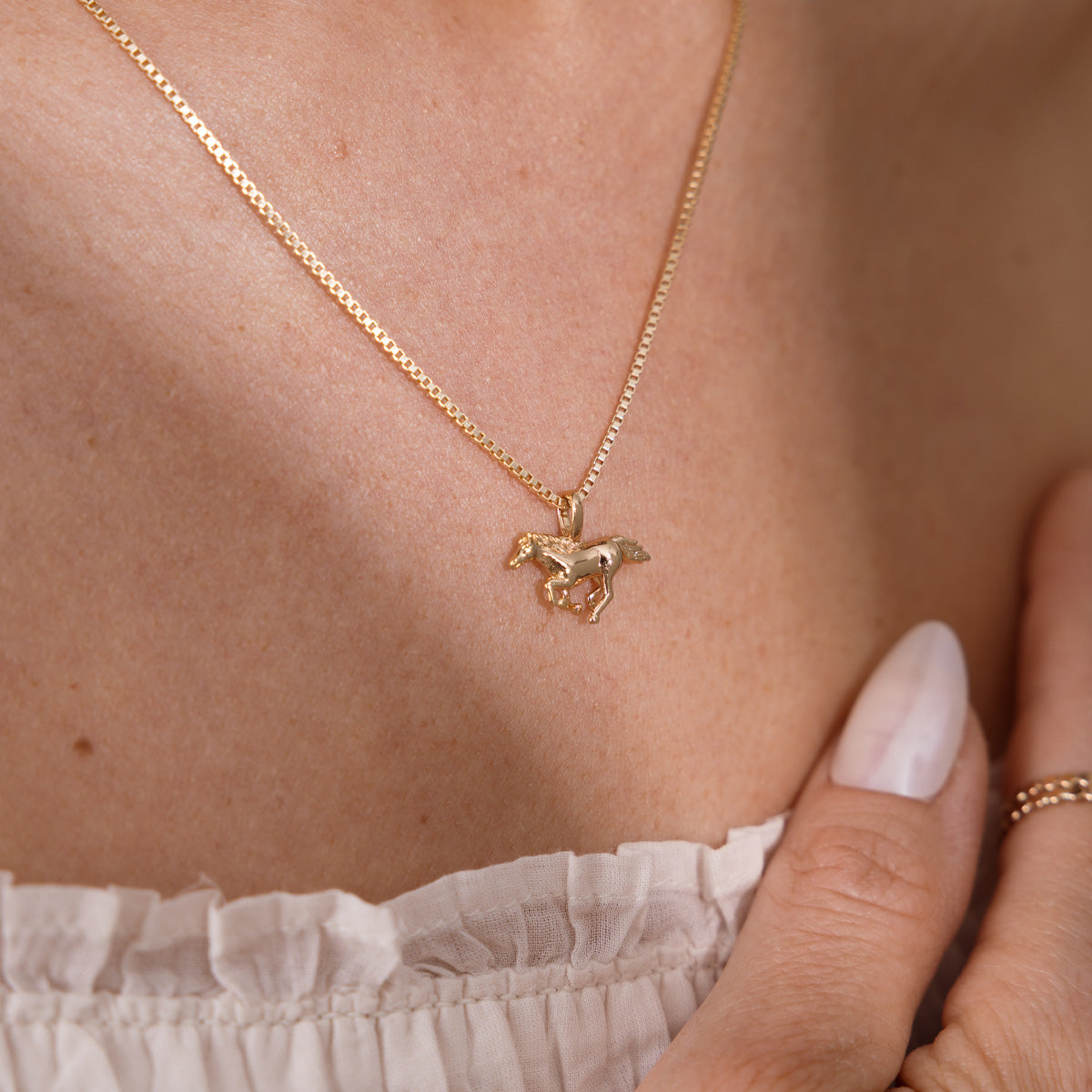 A close-up of a model wearing a delicate gold box chain necklace with a small horse pendant, styled with a white ruffled top and a gold ring.