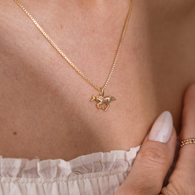 A close-up of a model wearing a delicate gold box chain necklace with a small horse pendant, styled with a white ruffled top and a gold ring.