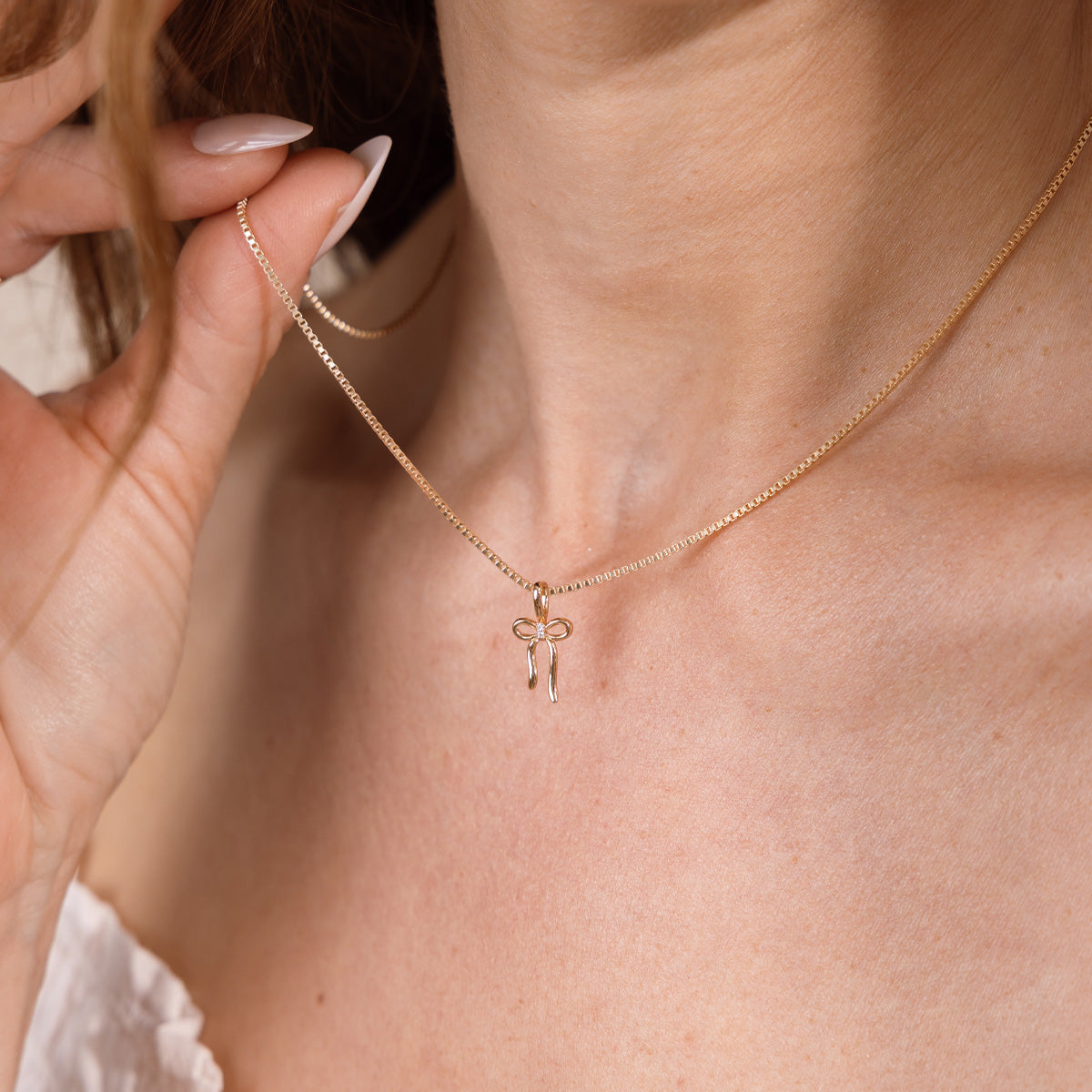 A close-up lifestyle shot of a model wearing a delicate gold chain necklace with a small bow-shaped pendant, held gently by a hand with manicured nails.
