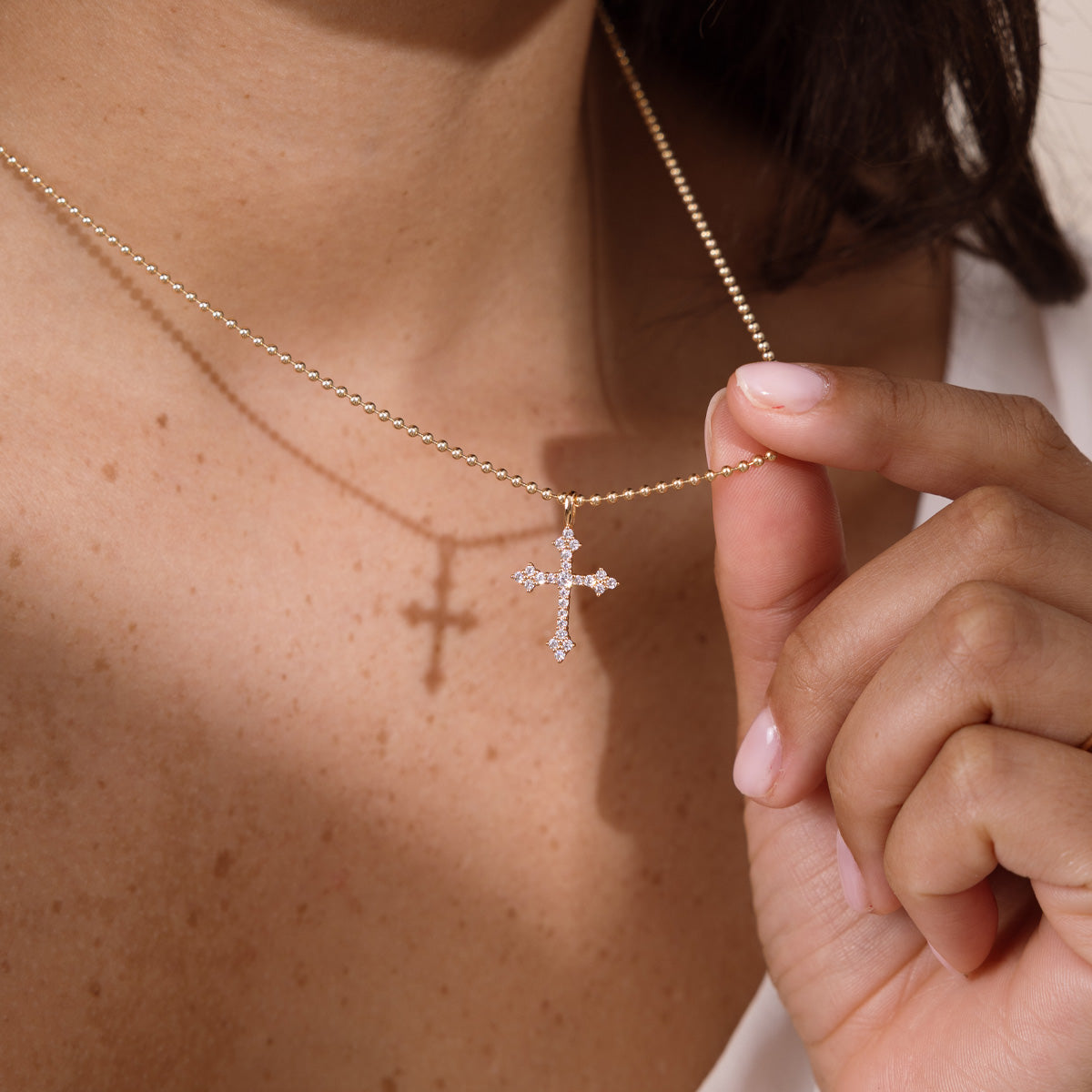 A close-up of a person wearing a delicate gold bead chain necklace with a small cross pendant adorned with clear stones, while a hand gently holds the chain.