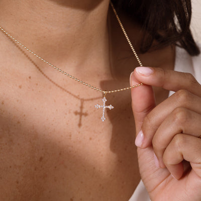 A close-up of a person wearing a delicate gold bead chain necklace with a small cross pendant adorned with clear stones, while a hand gently holds the chain.