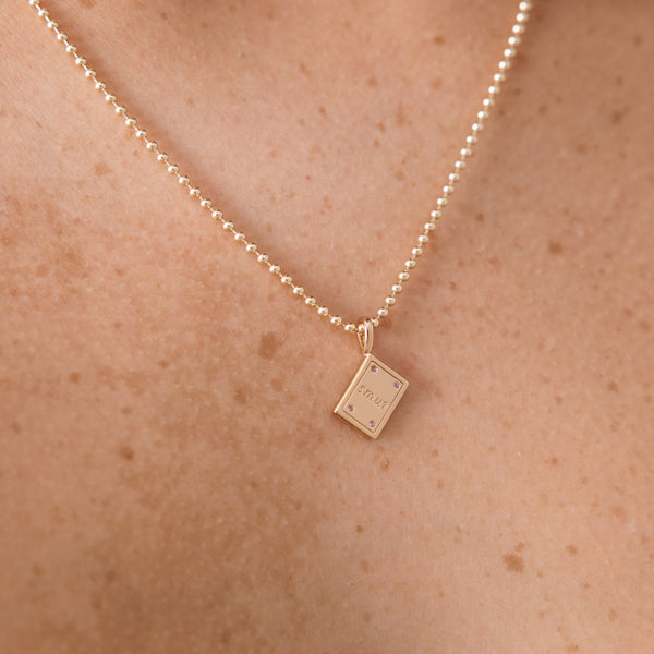 A delicate gold chain necklace with a small book pendant engraved with 'SMUT', worn on a person's neck against a warm skin tone background.