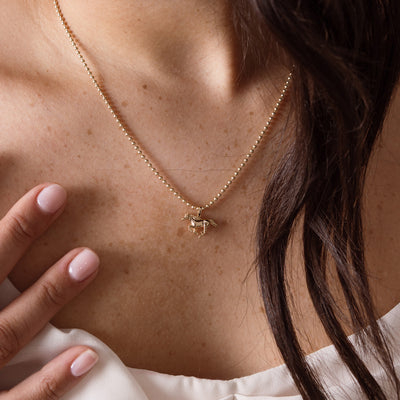 A close-up of a delicate gold bead chain necklace with a small horse pendant, worn on a model's neck with her hand resting near her chest.